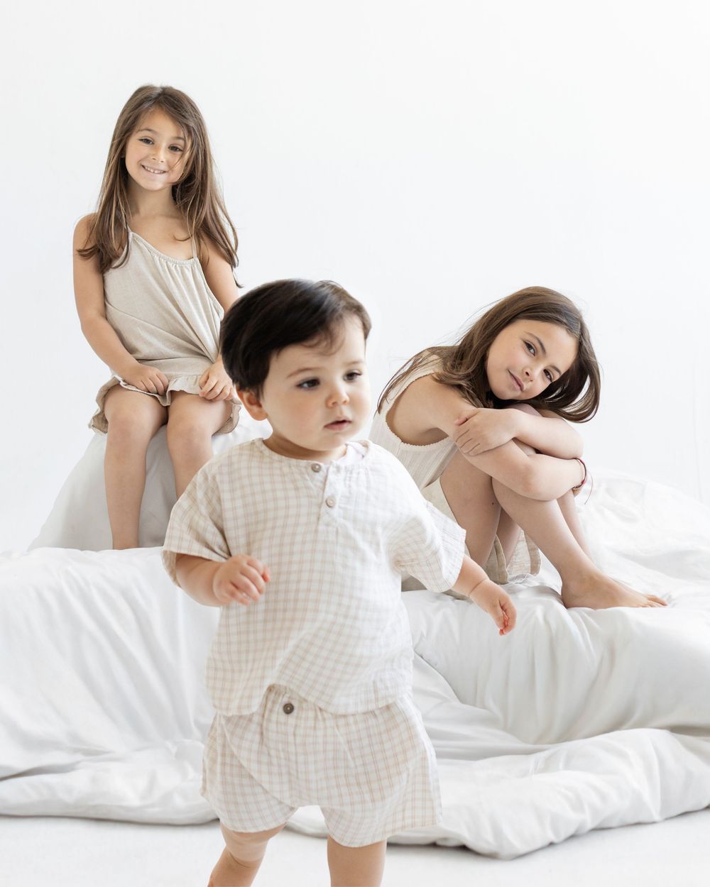 Three children in neutral linen outfits play on a white bed, with a toddler boy in a checkered set in the foreground.