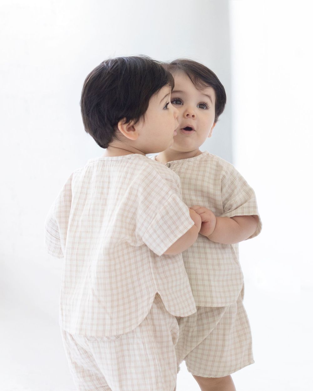 Baby toddler wearing matching beige gingham check short sleeve shirt and shorts set, reflected in mirror.