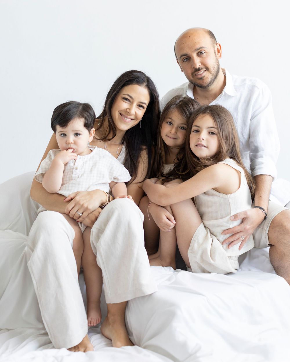 Happy family of five in white outfits sitting together on bed in bright minimalist studio portrait.