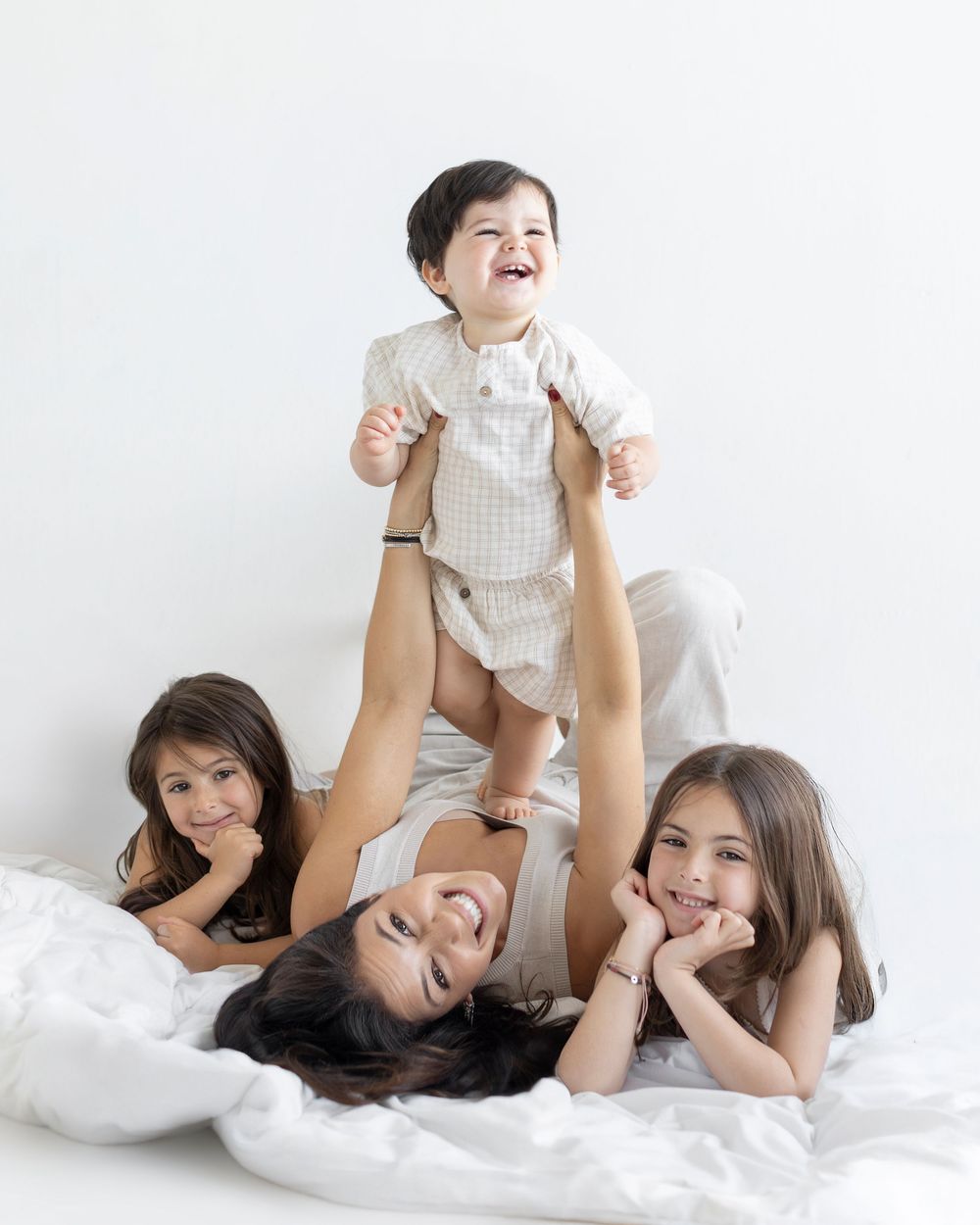 Mother lying down lifting laughing baby while two daughters smile in bright white studio photo.