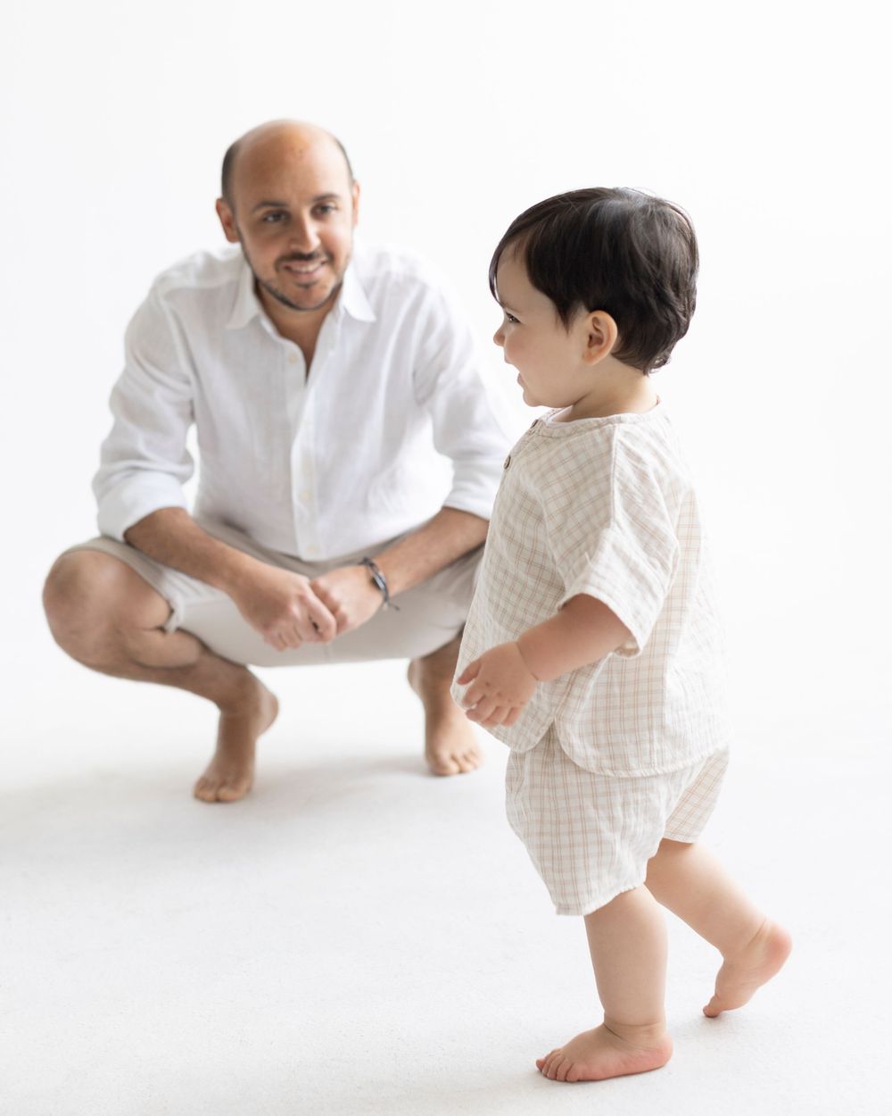 A bald man crouching in white shirt and shorts smiles at a toddler in checkered outfit learning to walk on white background.