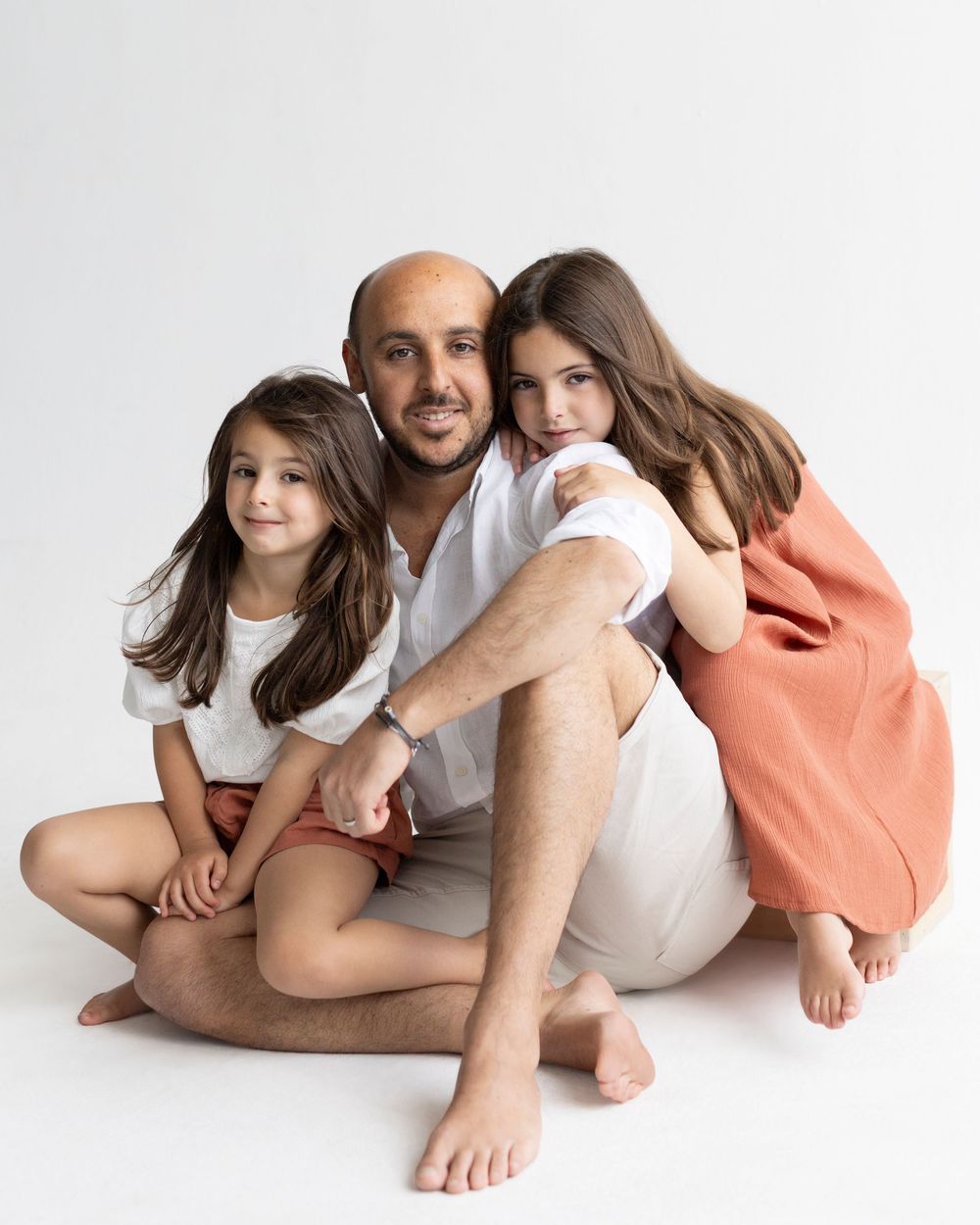 A bald man sits on white studio floor with two young girls with long brown hair, one in white, one in orange.