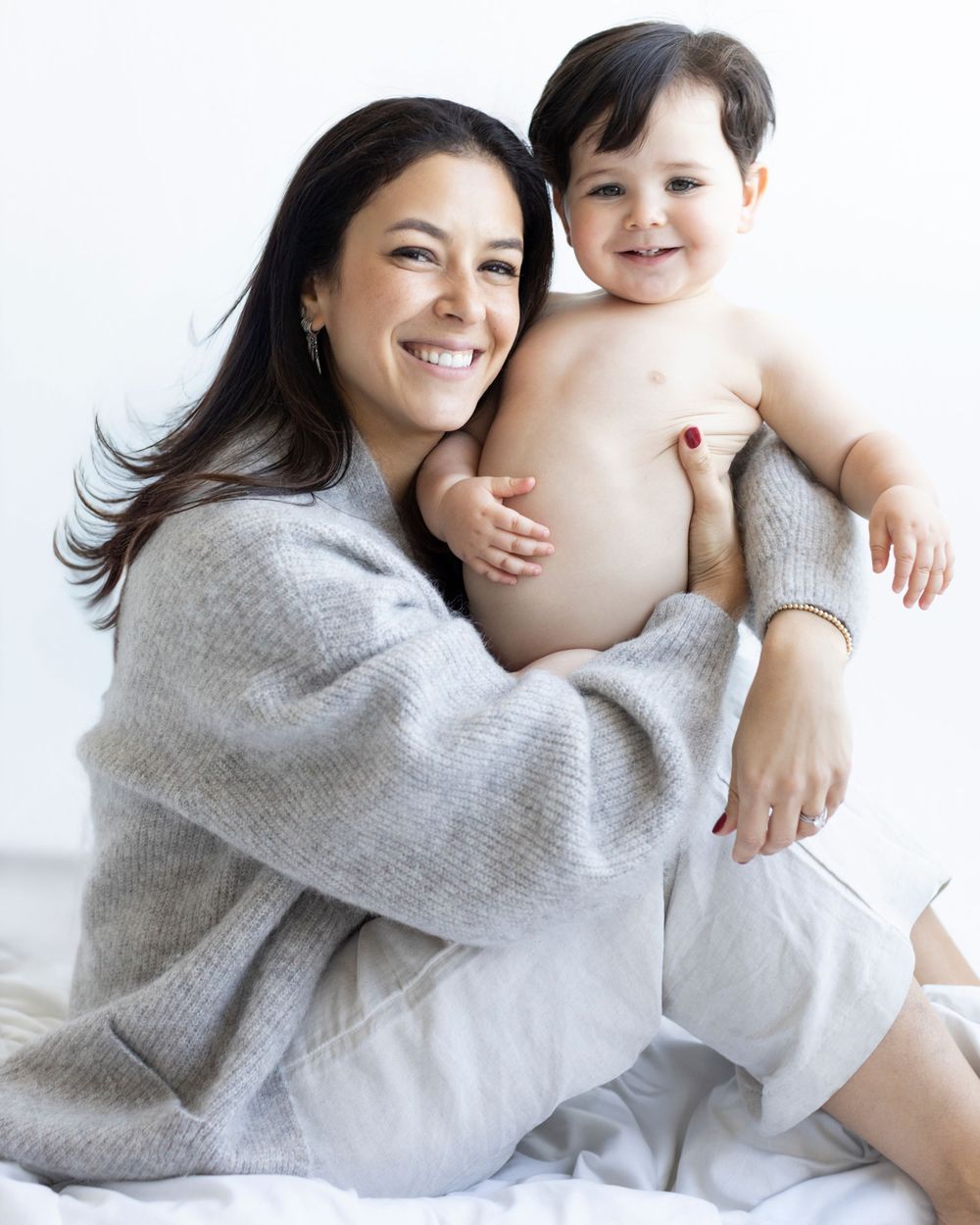 Smiling woman in gray sweater holding a happy shirtless baby on a white bed.