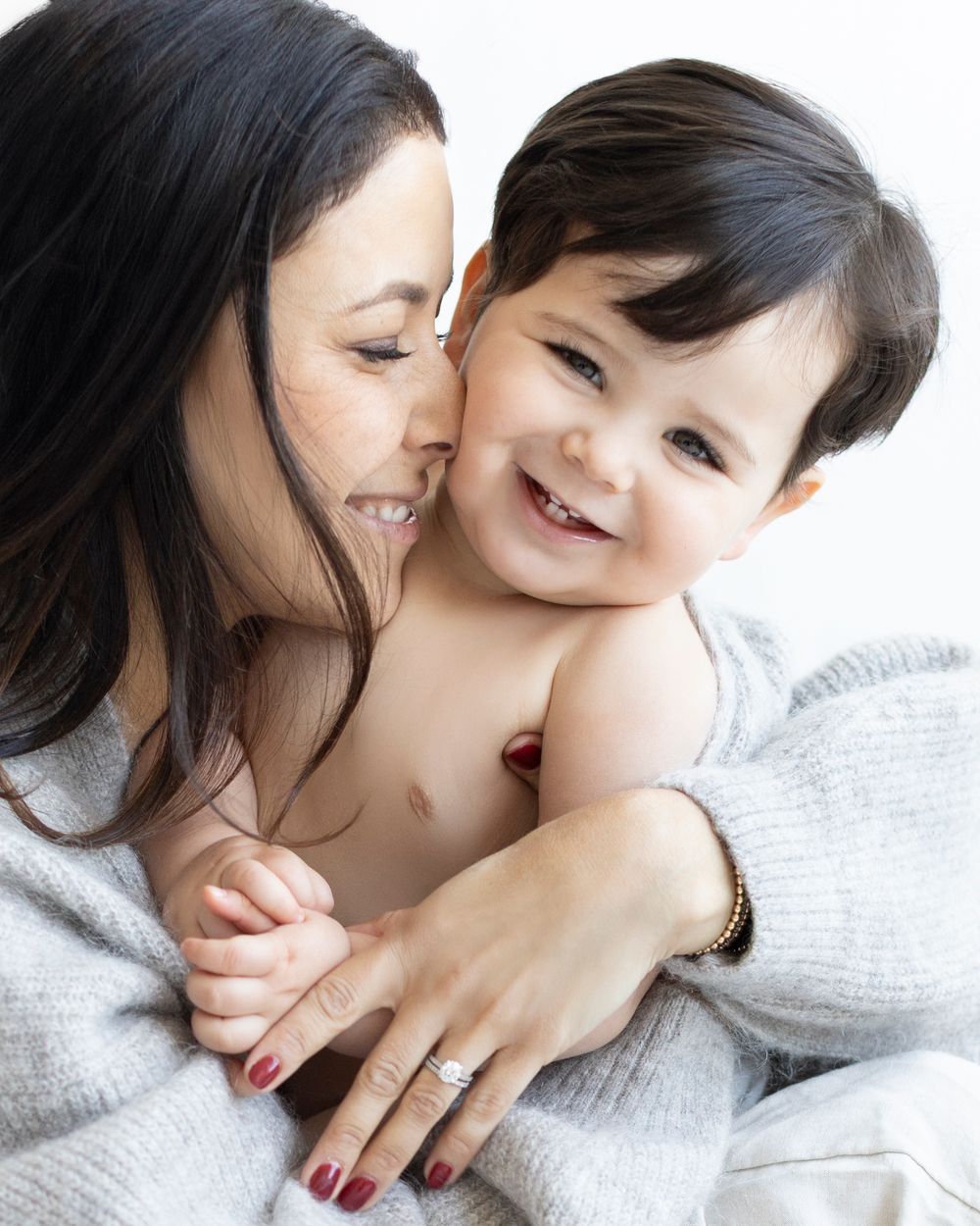 A smiling dark-haired woman in gray wrap holds a laughing baby boy with dark hair against white background.