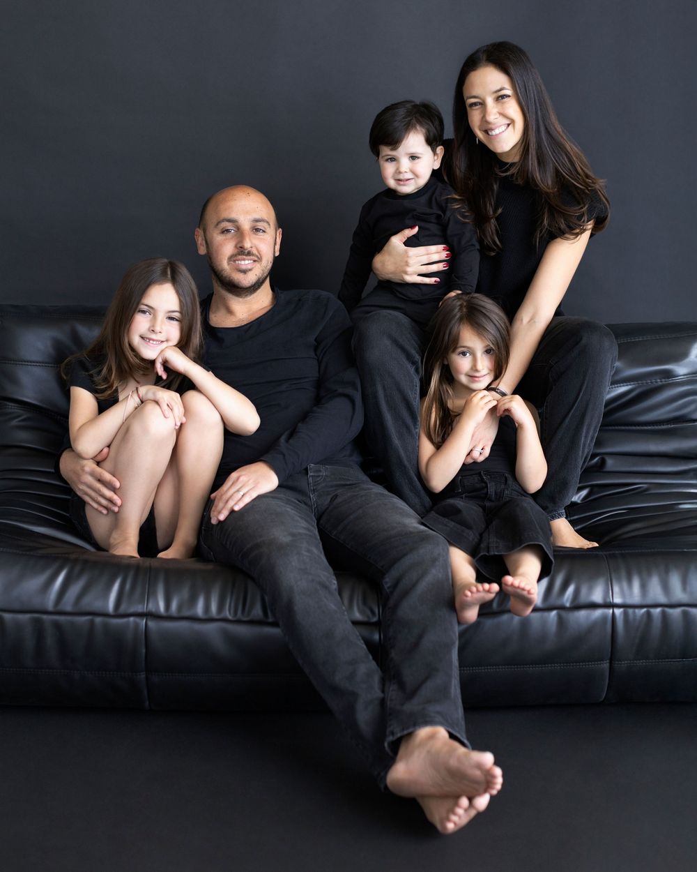 Family of five posing on black leather couch, all wearing matching black outfits against dark background.