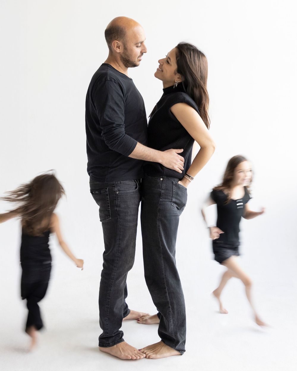 A couple embraces in a studio while two children run around them in motion blur, all dressed in black.