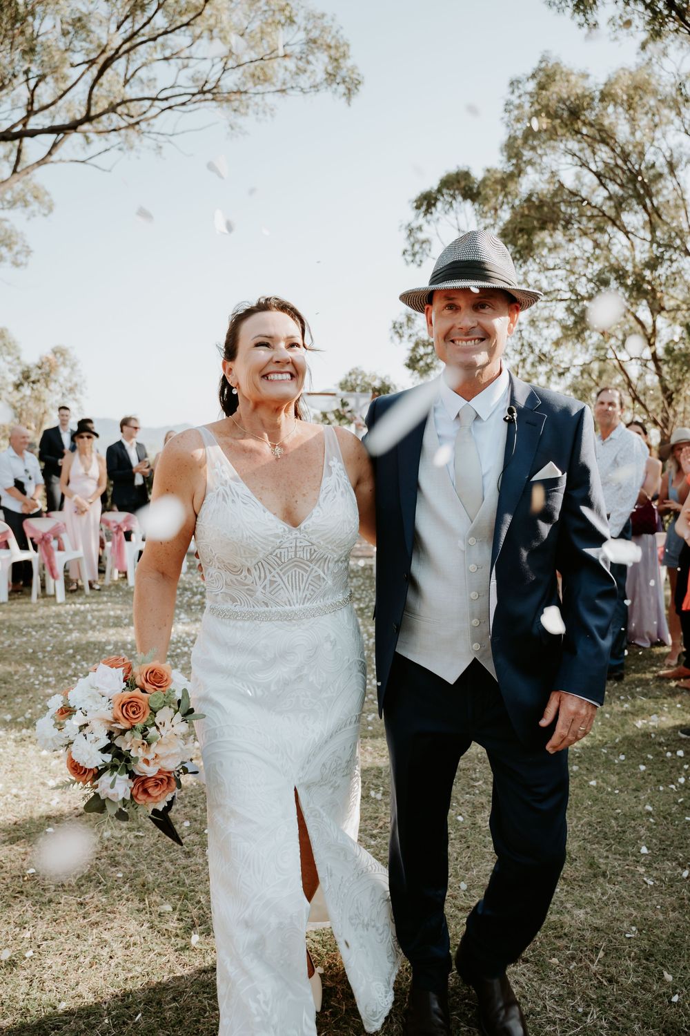 Wedding couple celebrates joyfully among trees at awoonga dam with guests in background.