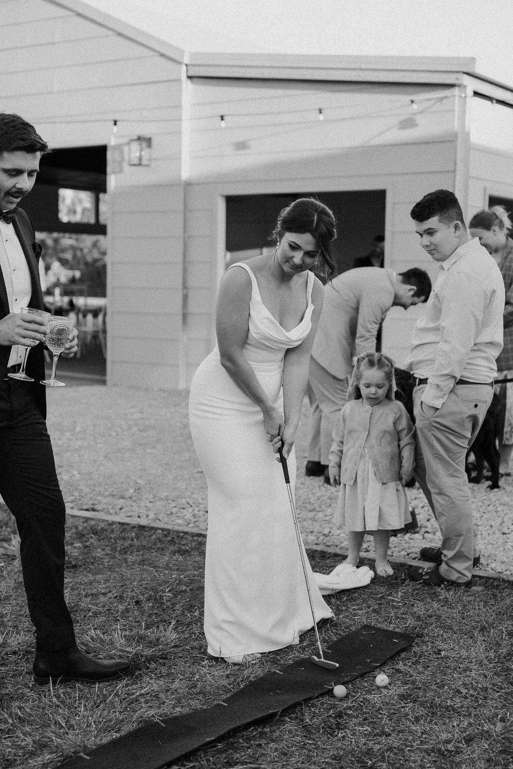 Bride in white gown playing mini golf with guests at outdoor wedding reception in black and white photo.