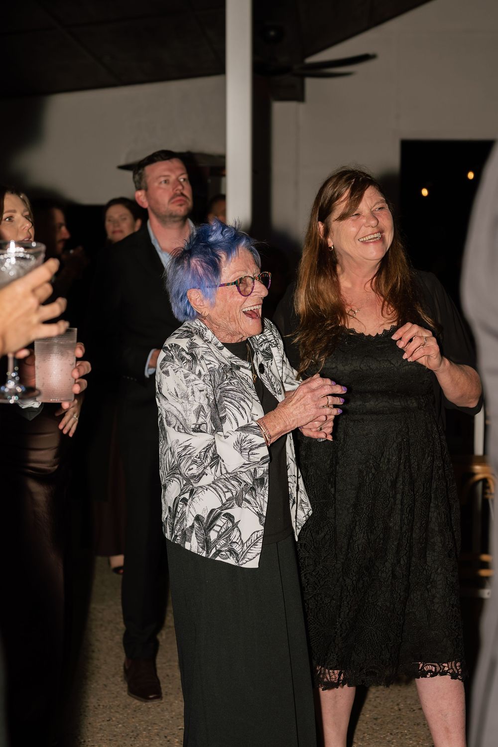 Two women laughing and dancing together at an evening event, one with blue hair wearing a floral jacket.