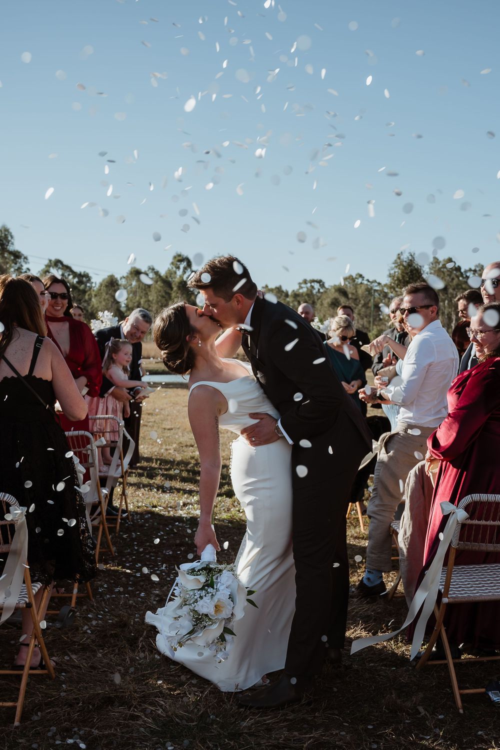 Bride and groom share a first kiss as newlyweds while white confetti fills the air at an outdoor wedding ceremony.