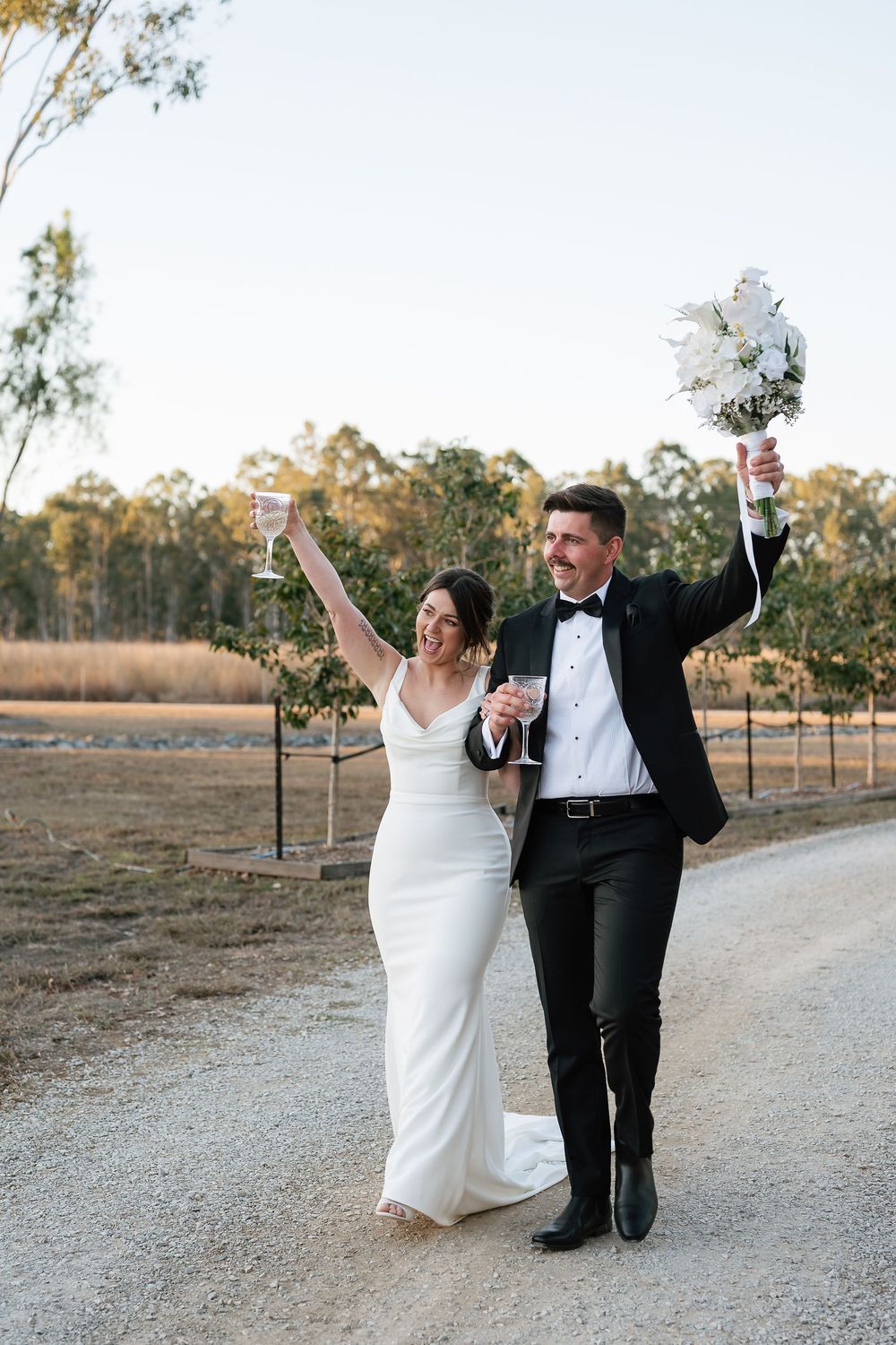 Joyful newlyweds celebrate on gravel path, bride in white gown holding bouquet, groom in black tuxedo, raising champagne glasses.