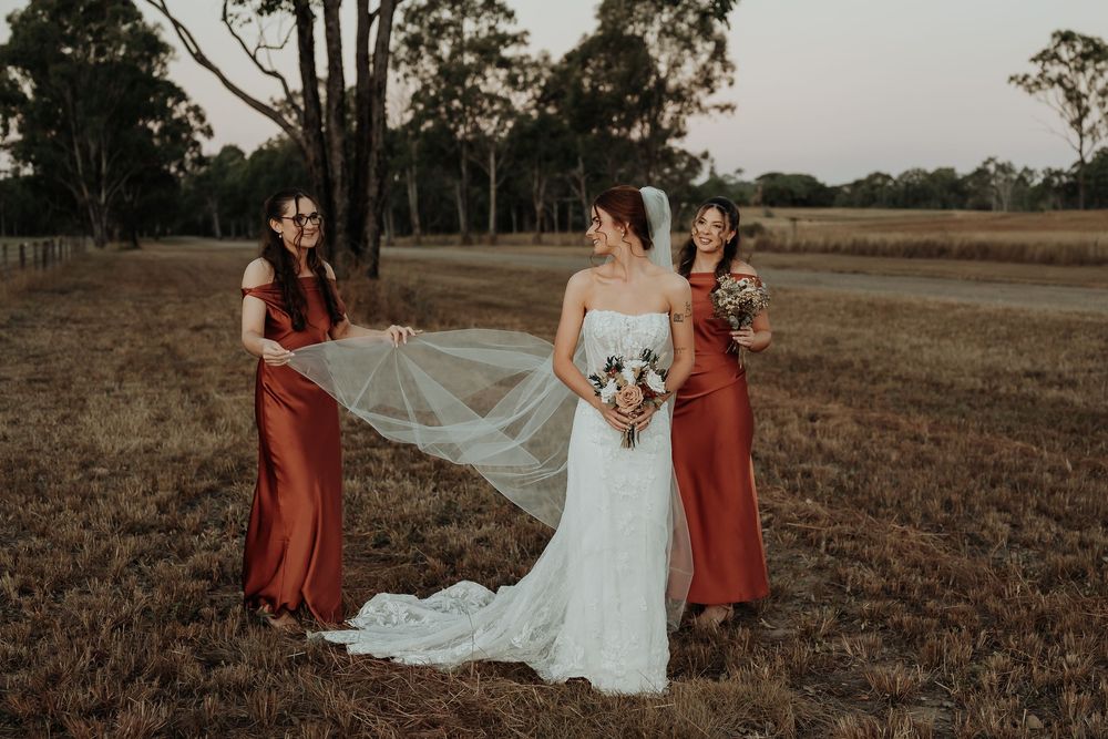 Bride in white gown with two bridesmaids in rust red dresses holding veil in a rustic field at sunset.