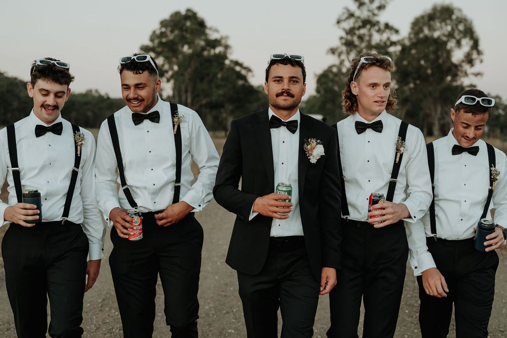 Five groomsmen in black tuxedos with bow ties and suspenders walk together outdoors holding drinks at a wedding.