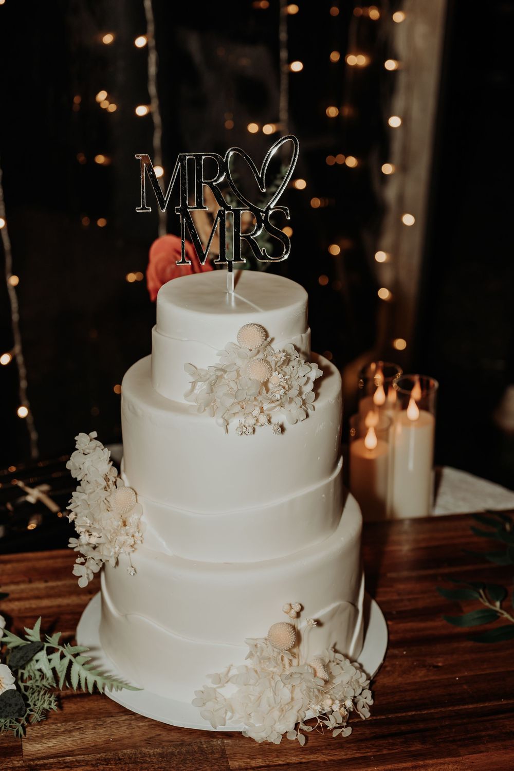 Elegant four-tier white wedding cake with floral accents and Mr & Mrs cake topper, displayed on a wooden table with candles.