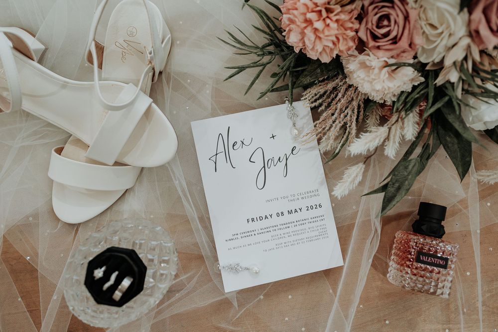 Elegant wedding flat lay with white heels, floral bouquet, invitation, perfume, and earrings on sheer fabric.