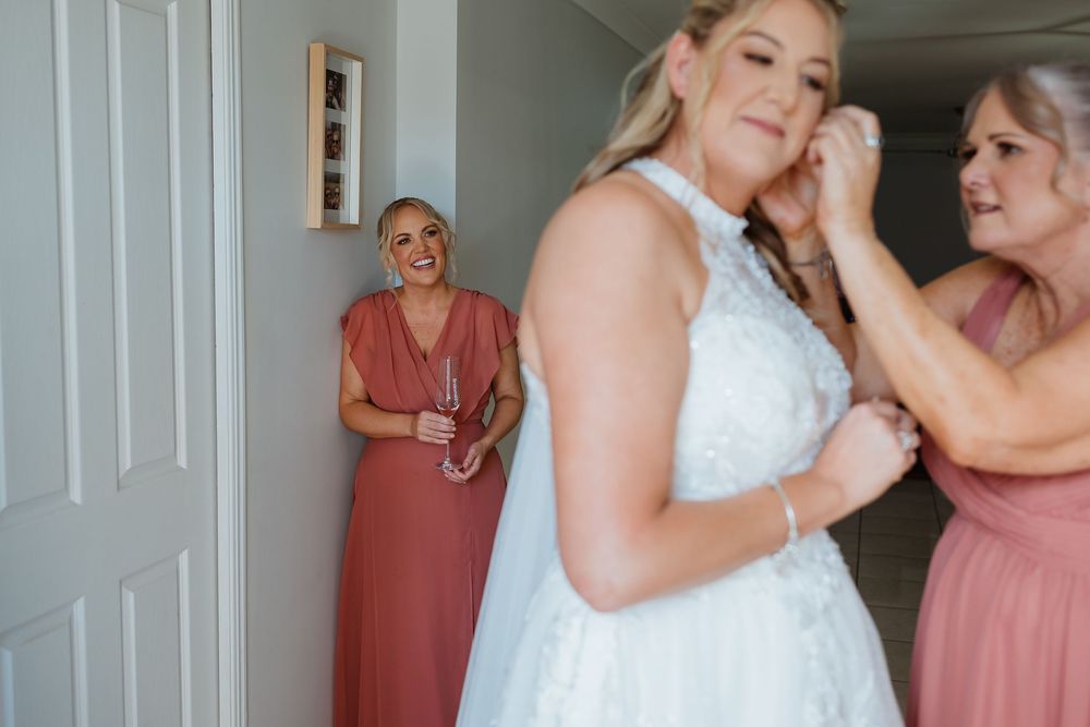 Bride getting dressed with bridesmaids in pink helping fasten her white lace wedding gown before the ceremony.