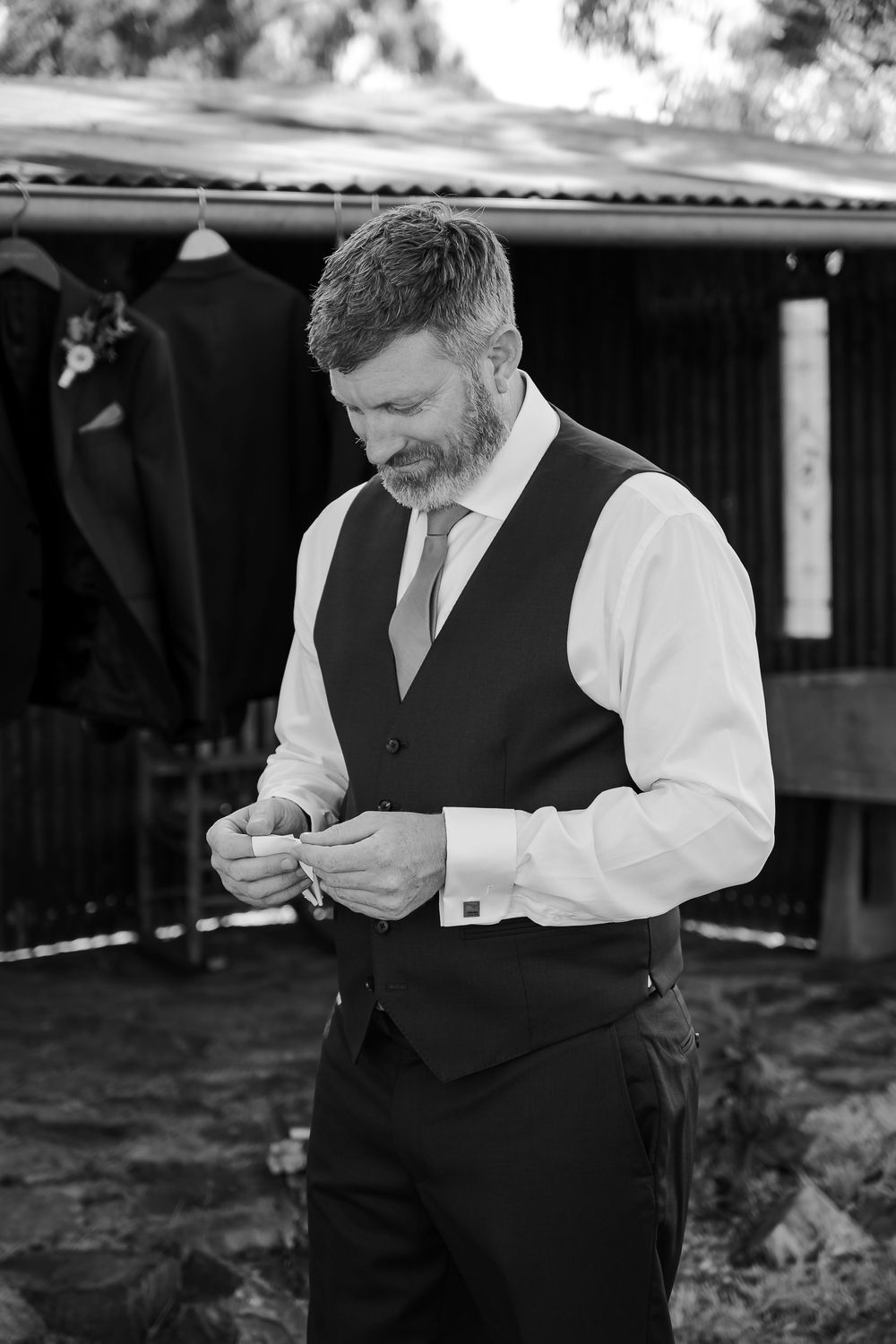 Black and white photo of a bearded groom in a vest and tie fastening his cufflinks outdoors before his wedding.
