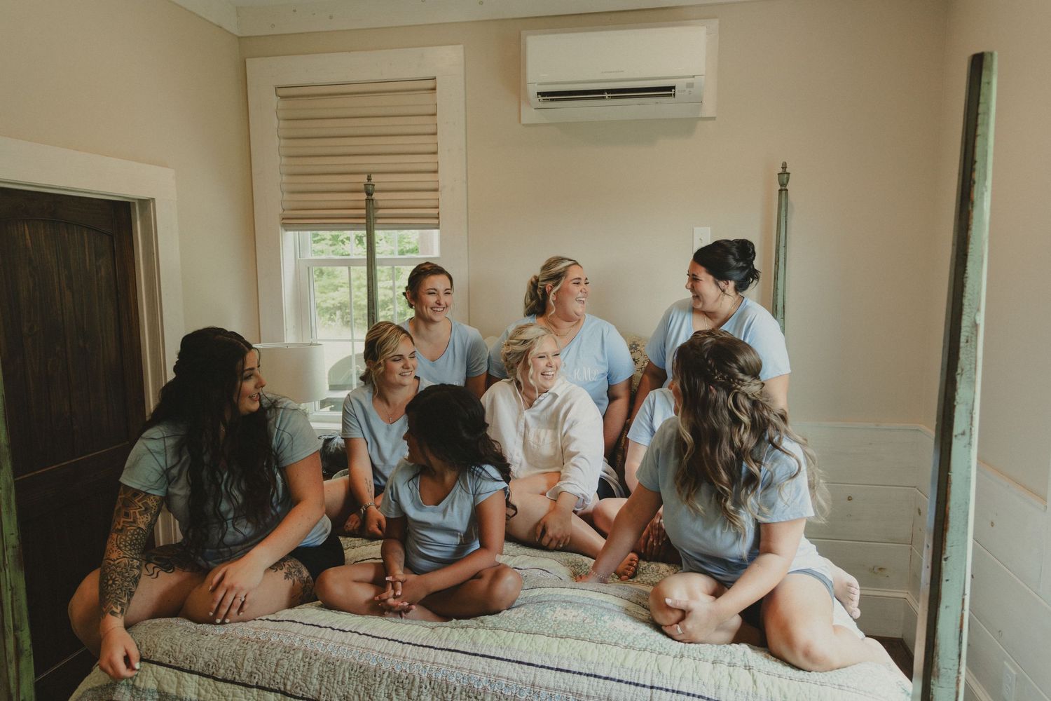 A group relaxes on a bed in casual attire during what appears to be a pre-wedding gathering.