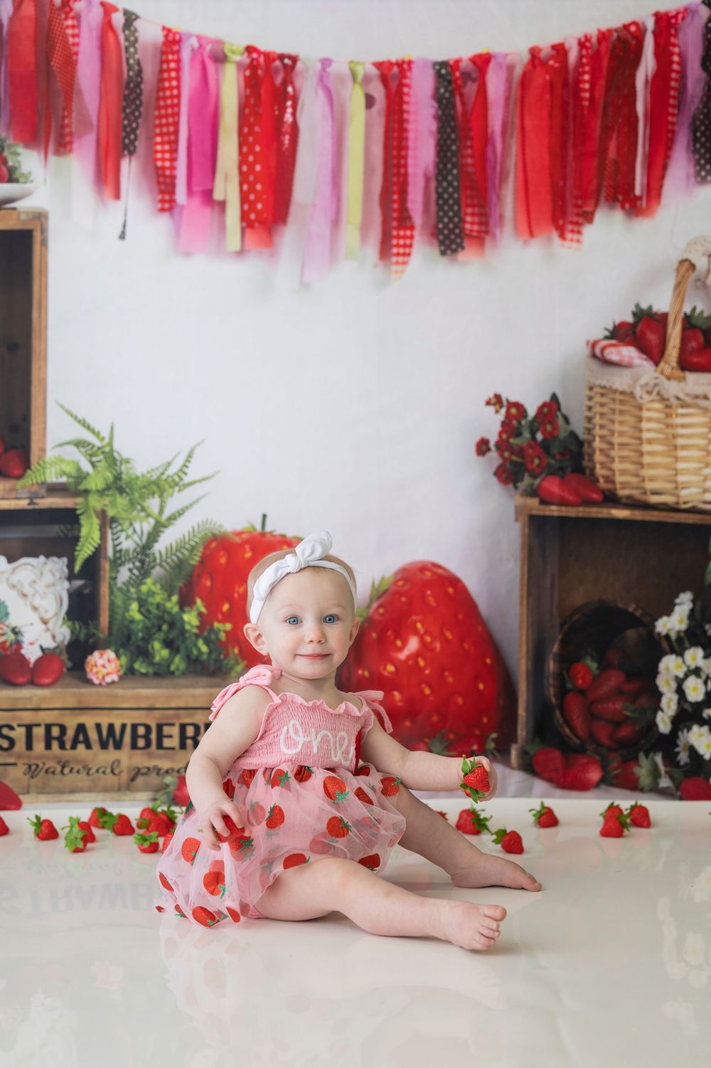 Strawberry-themed baby photo session with red and pink ribbon banner decorations and rustic backdrop.