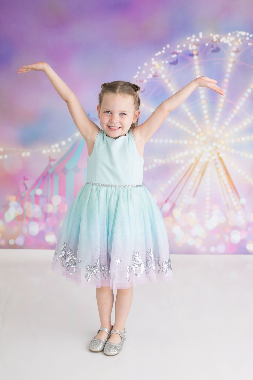 A young child in a mint blue tulle dress poses with arms raised against a magical carnival background with twinkling lights.