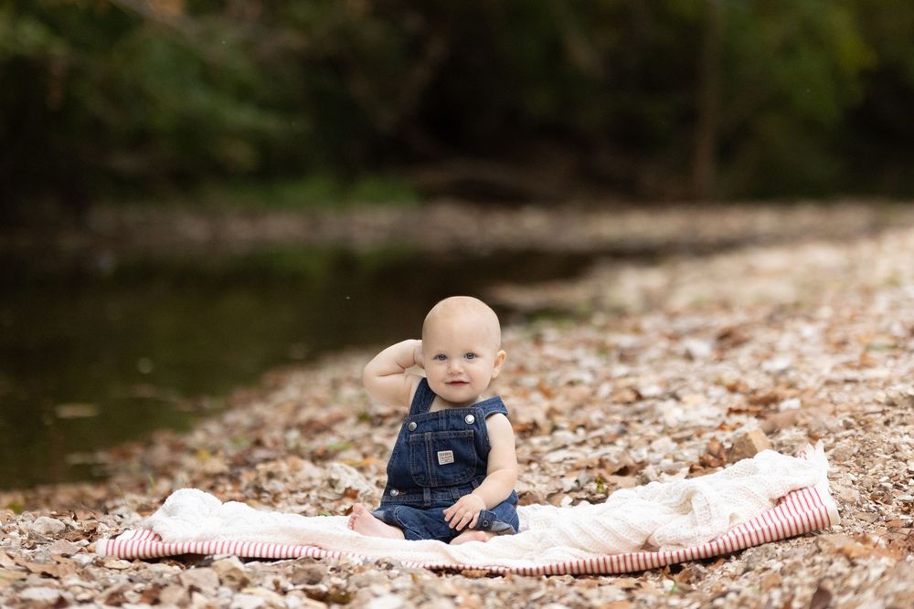 Baby in blue overalls sits on a blanket by the water's edge on a rocky shore.