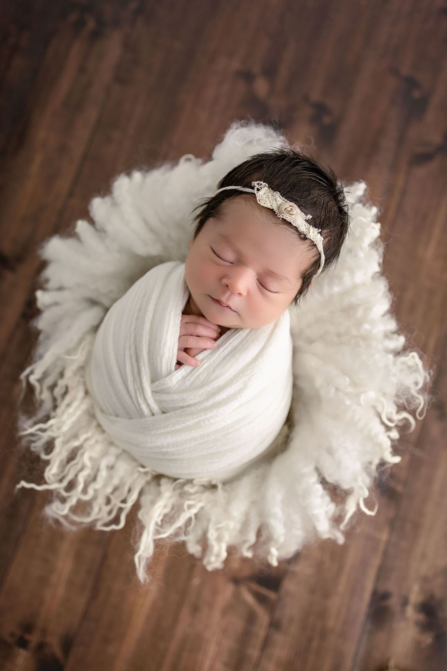 Sleeping newborn wrapped in white cloth nestled on soft fluffy white blanket against wooden floor.