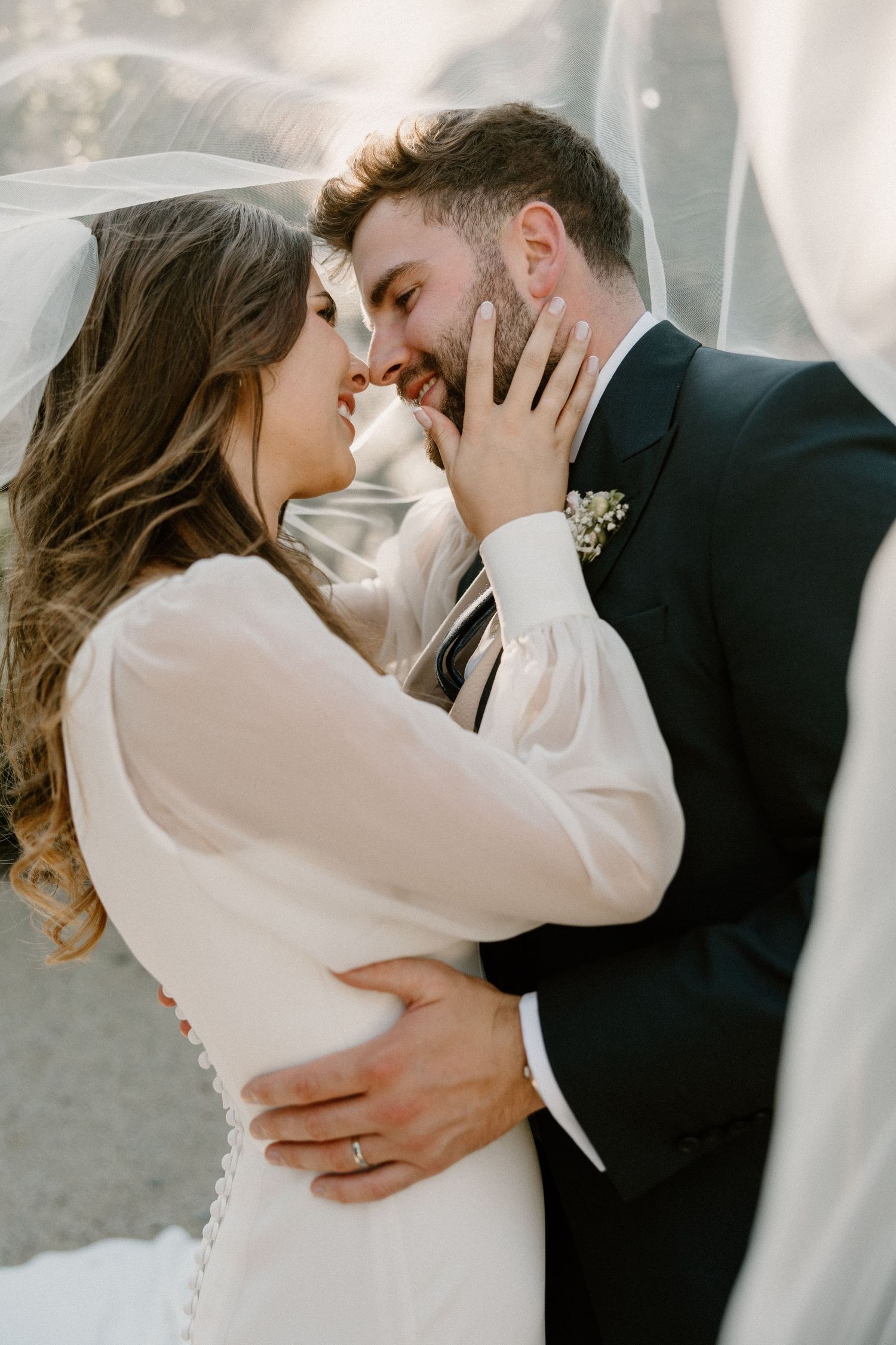 A couple in wedding attire share an intimate moment under a flowing veil during their wedding ceremony.