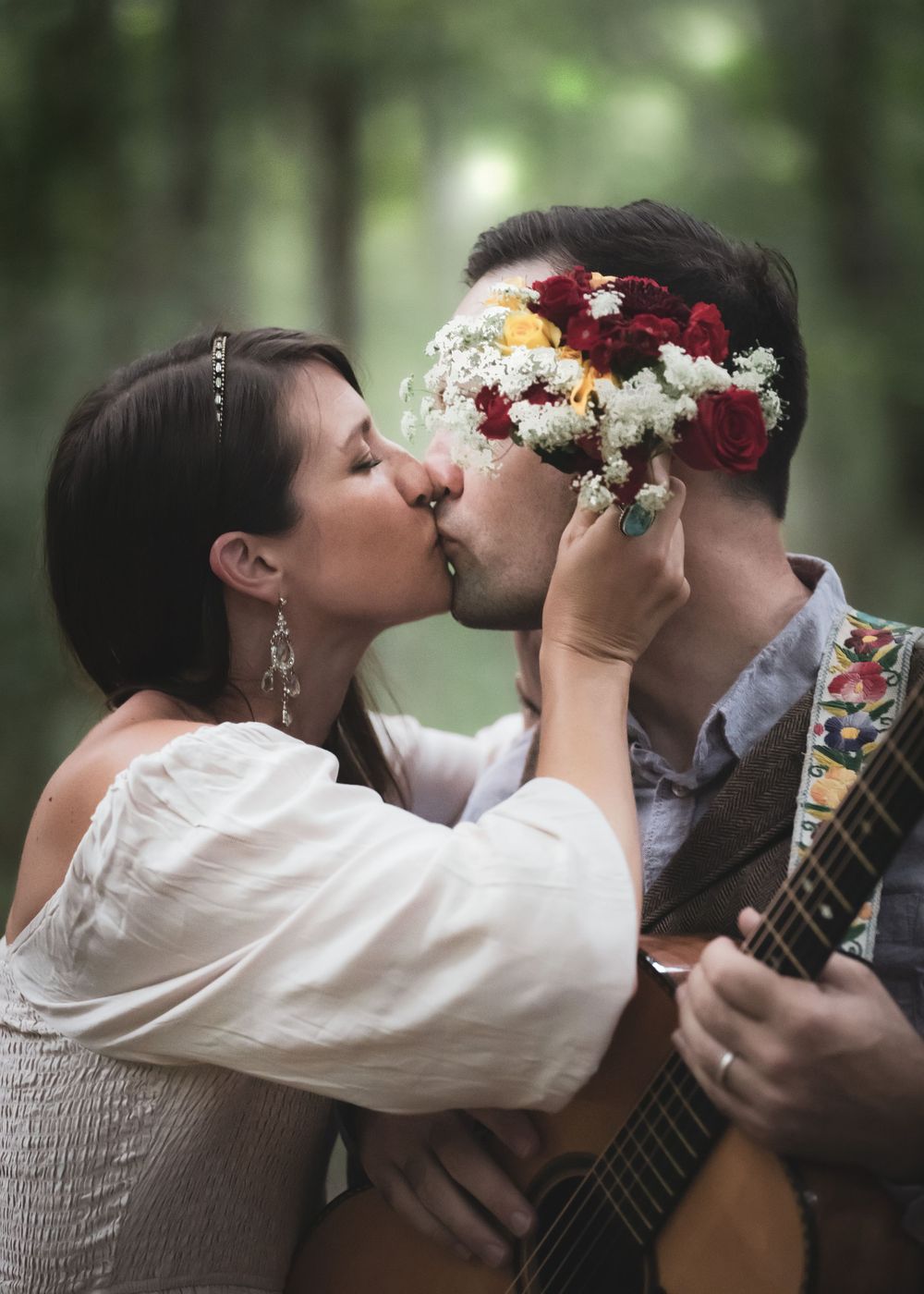 Couple sharing a romantic kiss in a forest, with the groom playing guitar and bride holding a colorful floral bouquet.