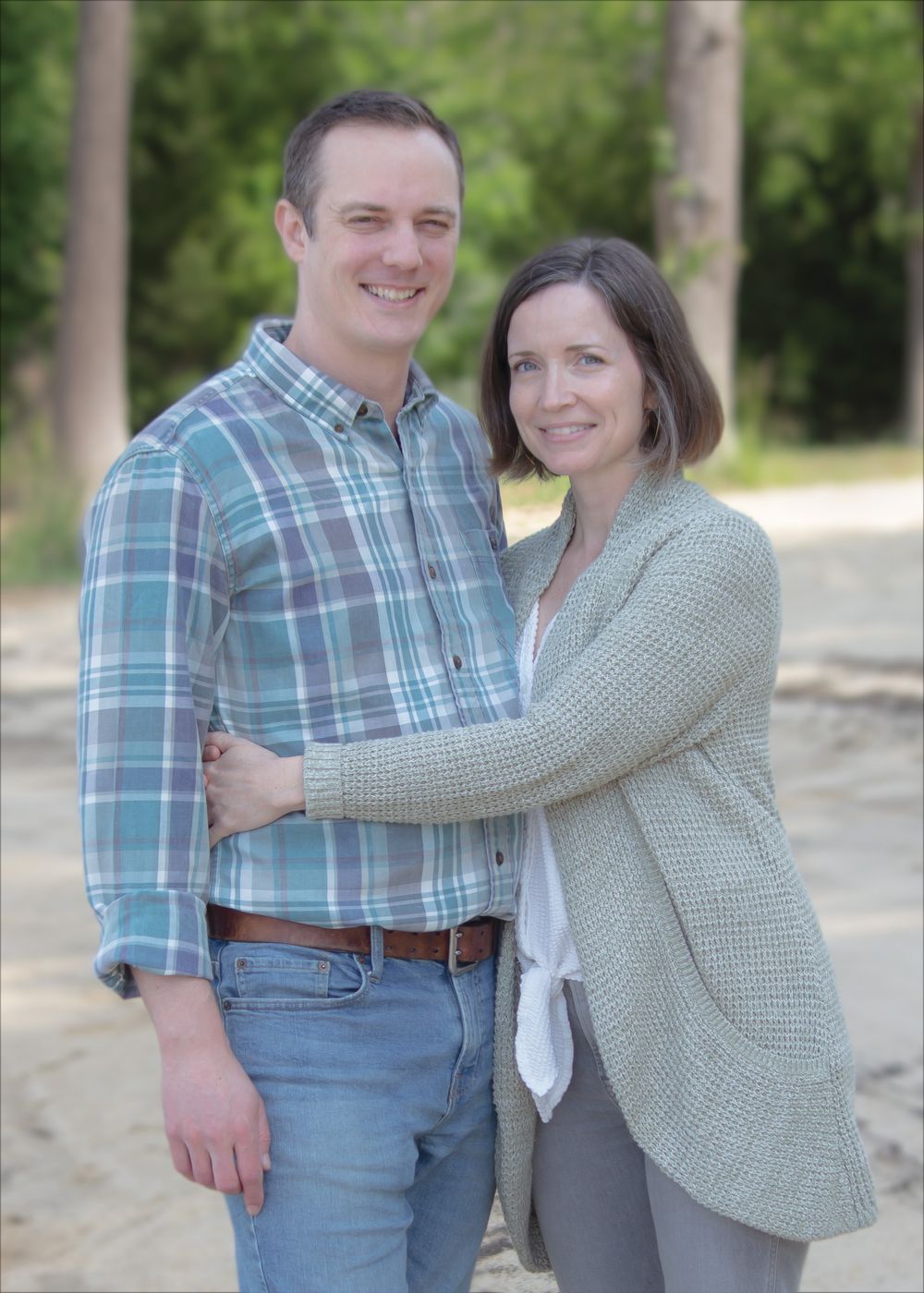 Smiling couple embracing outdoors on sandy path surrounded by green trees, man in plaid shirt and woman in sage cardigan.