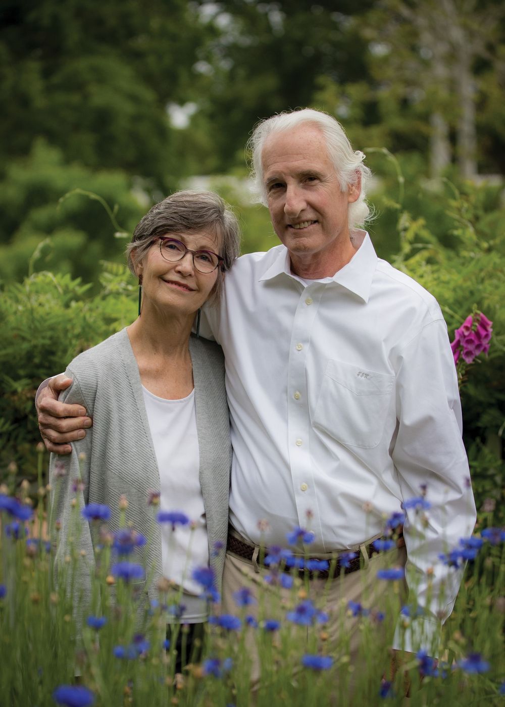 A smiling senior couple stands embracing in a vibrant garden filled with blue cornflowers and greenery.