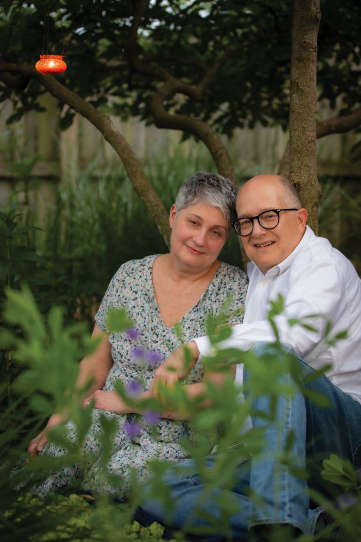 A happy senior couple sits together among lush garden greenery, smiling warmly at the camera.