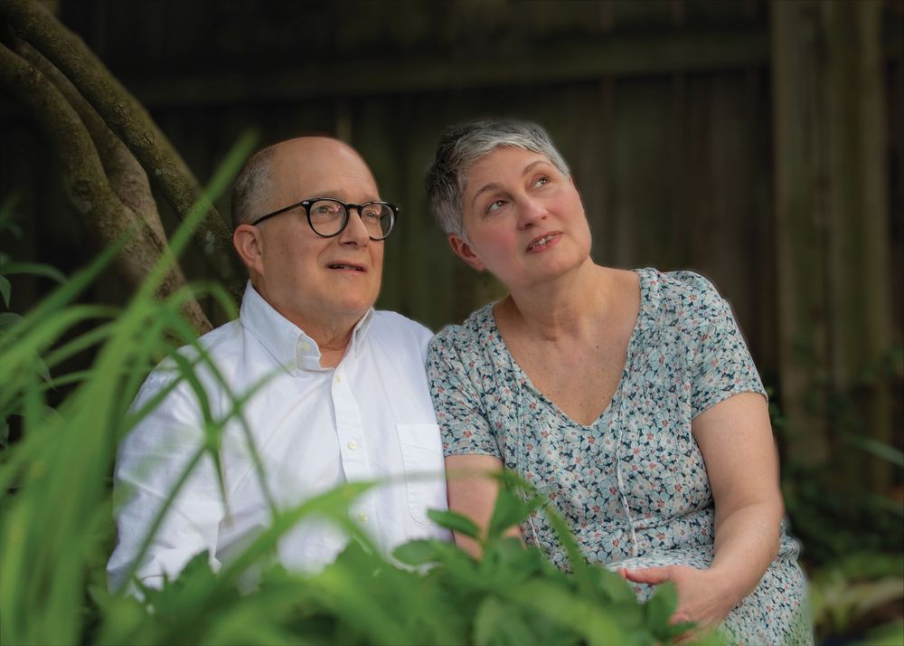 An older couple sits peacefully in a garden, the man in glasses gazing upward while the woman leans close.