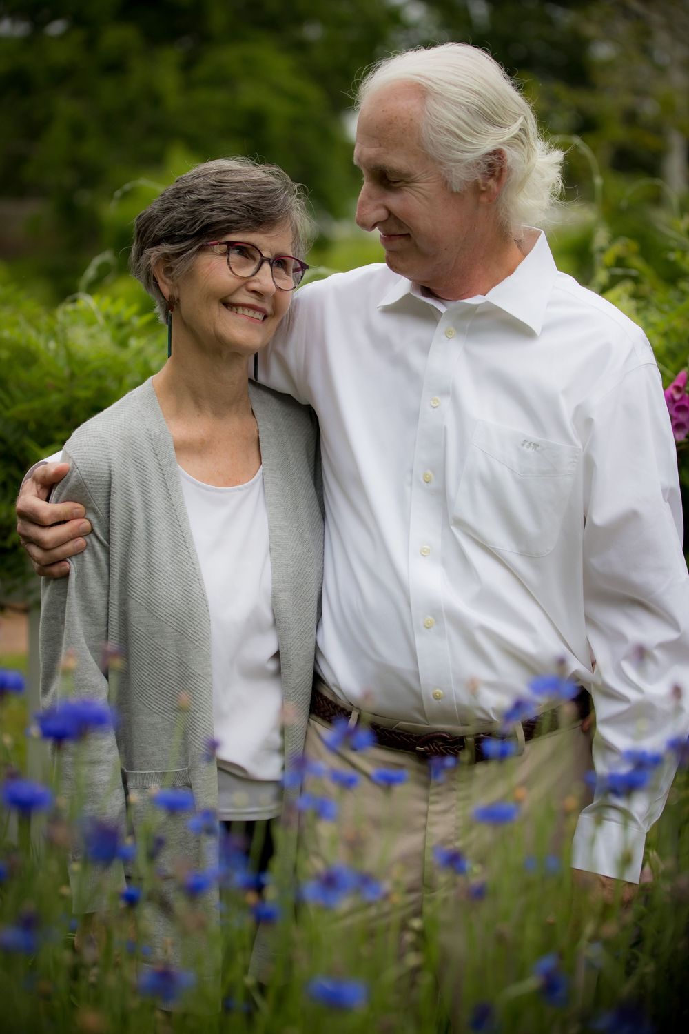 An older couple stands arm in arm among blue wildflowers, the man looking lovingly at the woman.