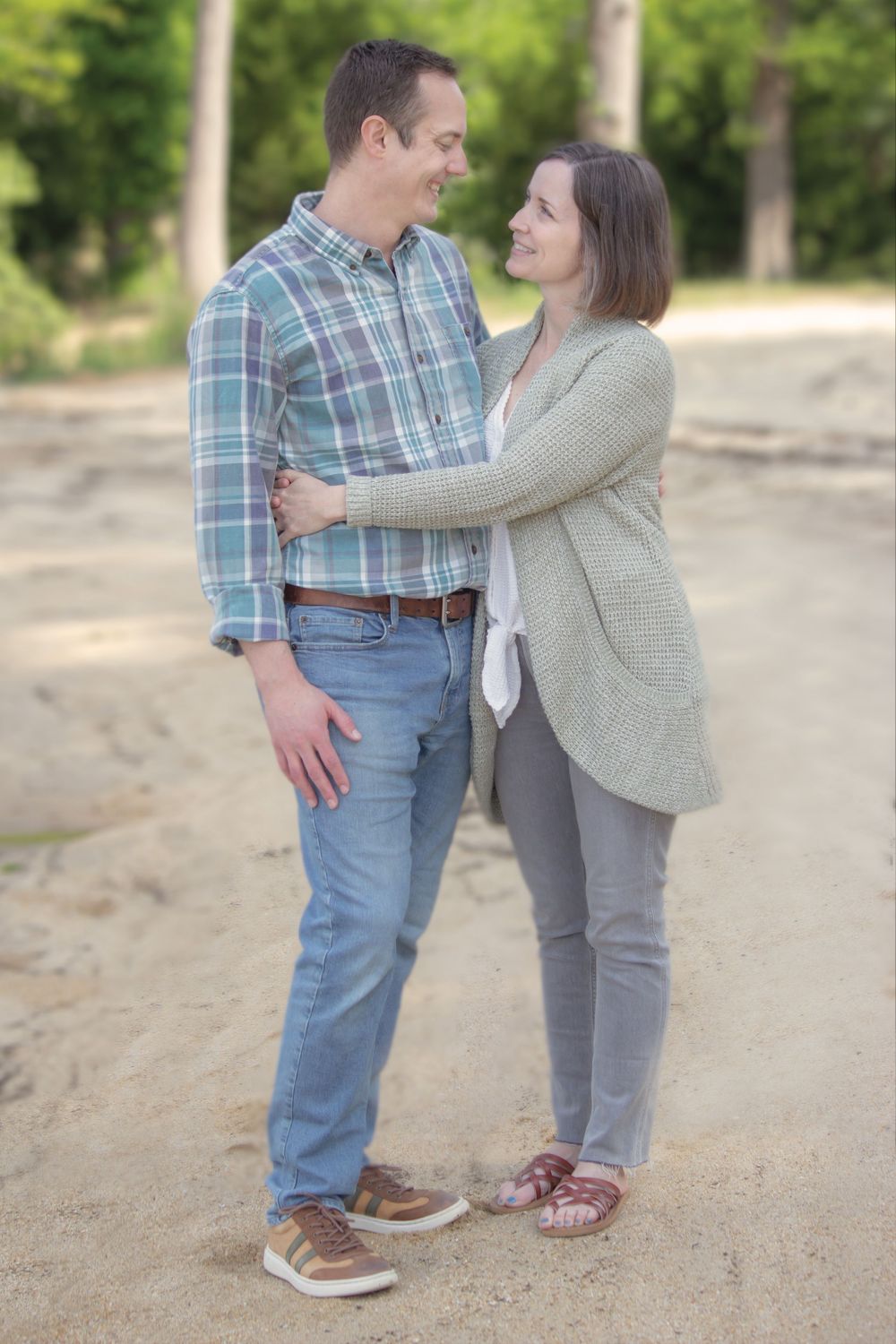 Happy couple holding each other and gazing into each others eyes on a sandy outdoor path with trees in background.