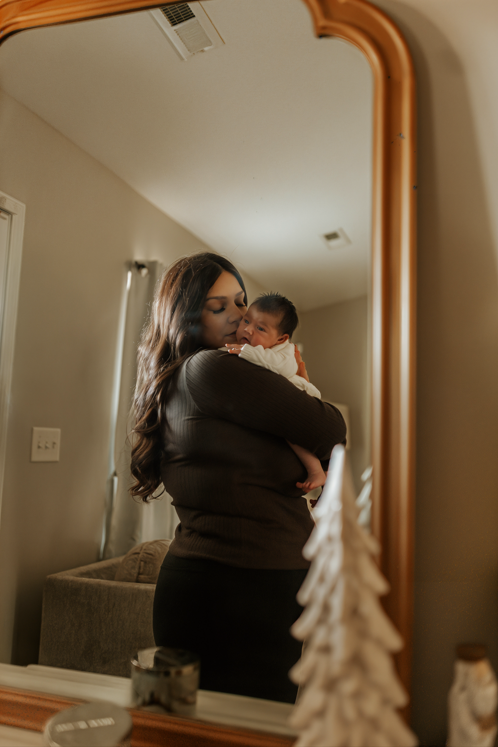 Mirror reflection shows a tender moment between parent and baby in a cozy room with decorative holiday trees.