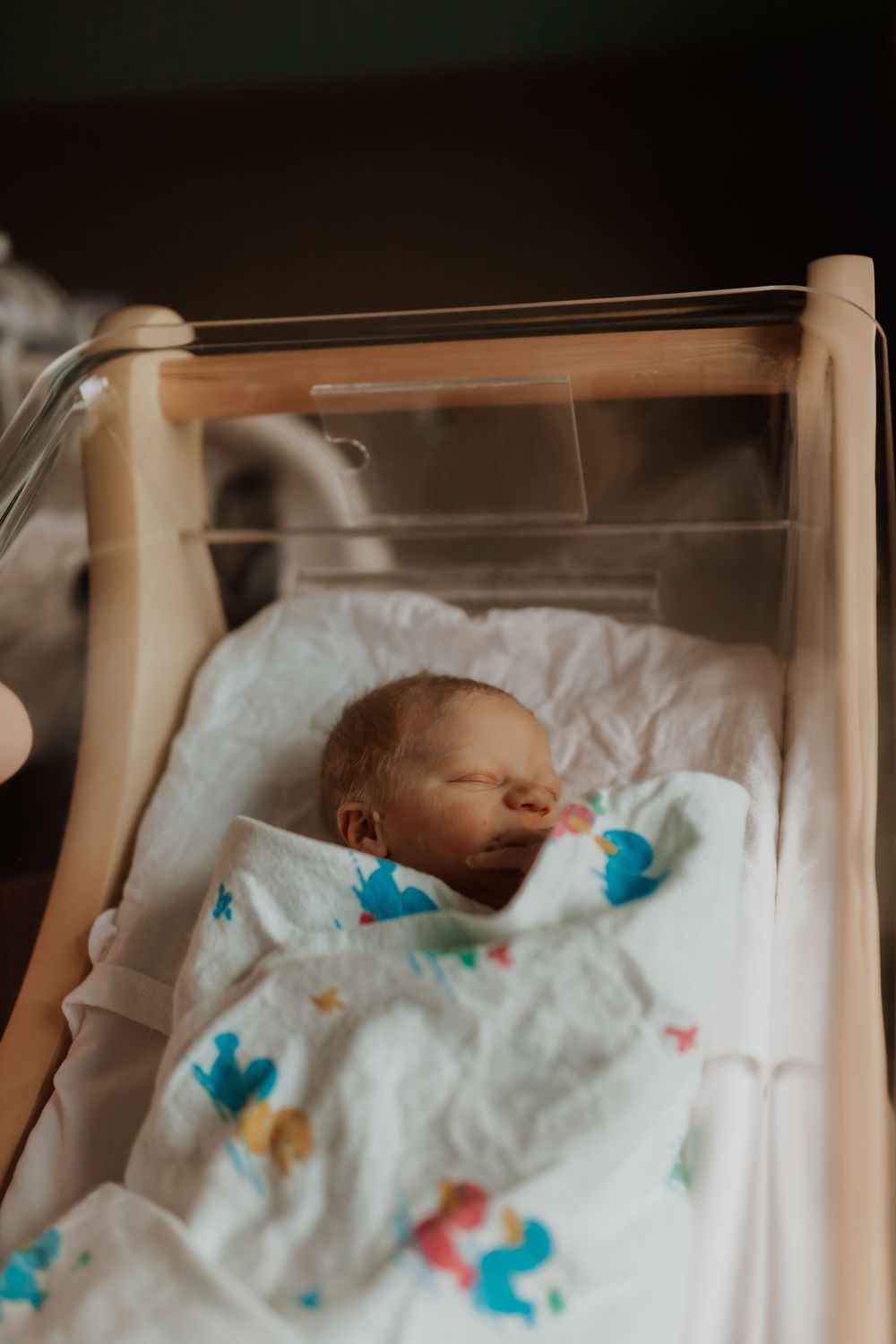 Newborn baby sleeping peacefully in hospital bassinet with colorful patterned blanket.