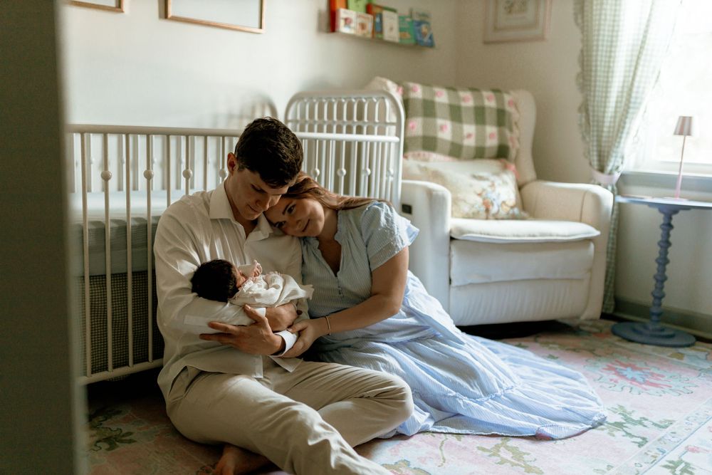 New parents sitting on nursery floor holding their newborn baby together near a white crib.