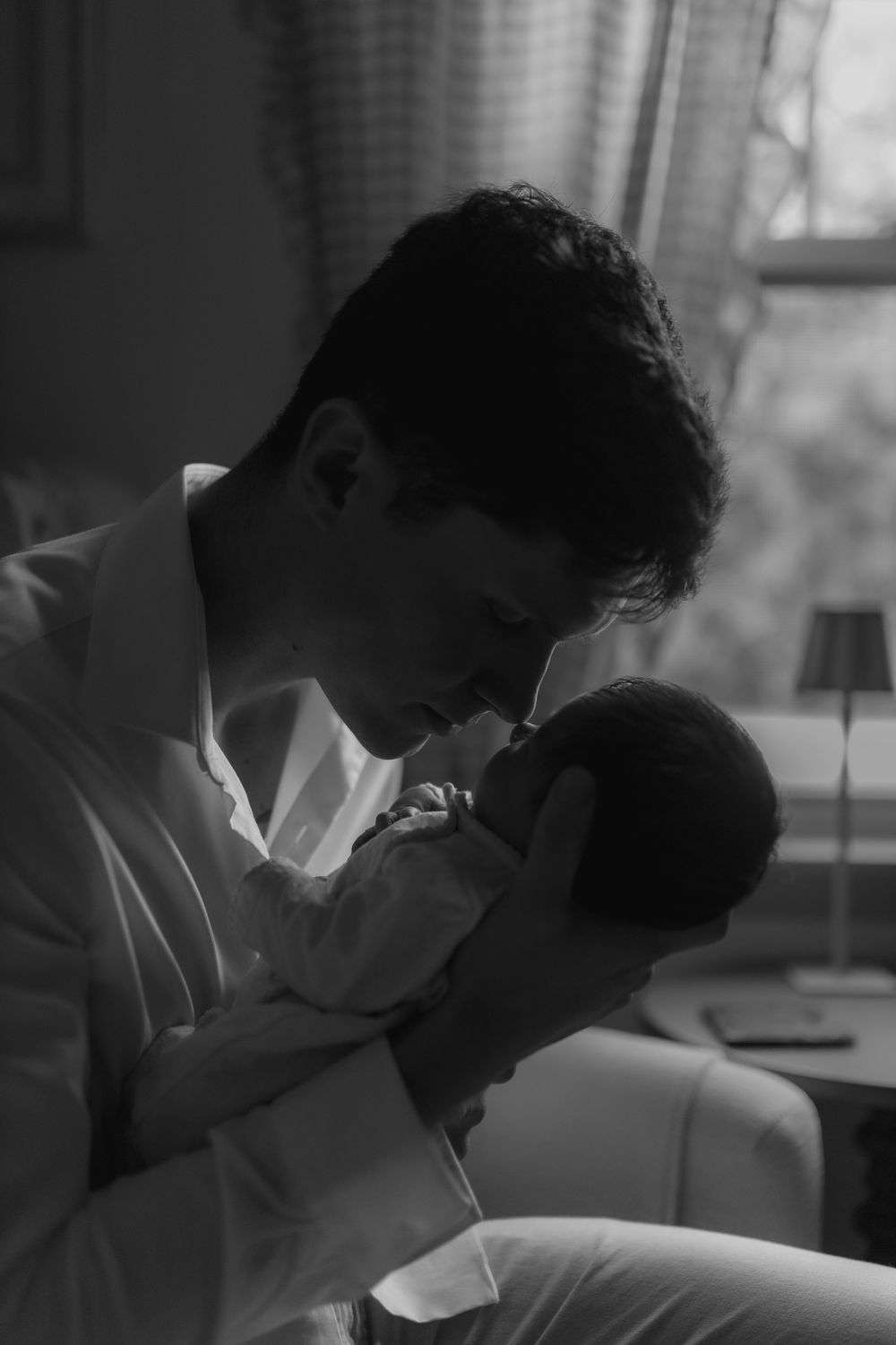 Black and white photo of a father tenderly holding and bonding with his newborn baby near a window.