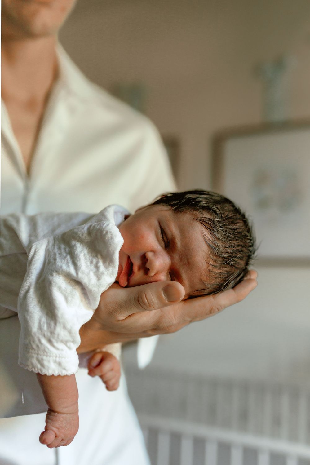 A parent in white gently cradles a sleeping newborn baby in one hand inside a softly lit nursery room.
