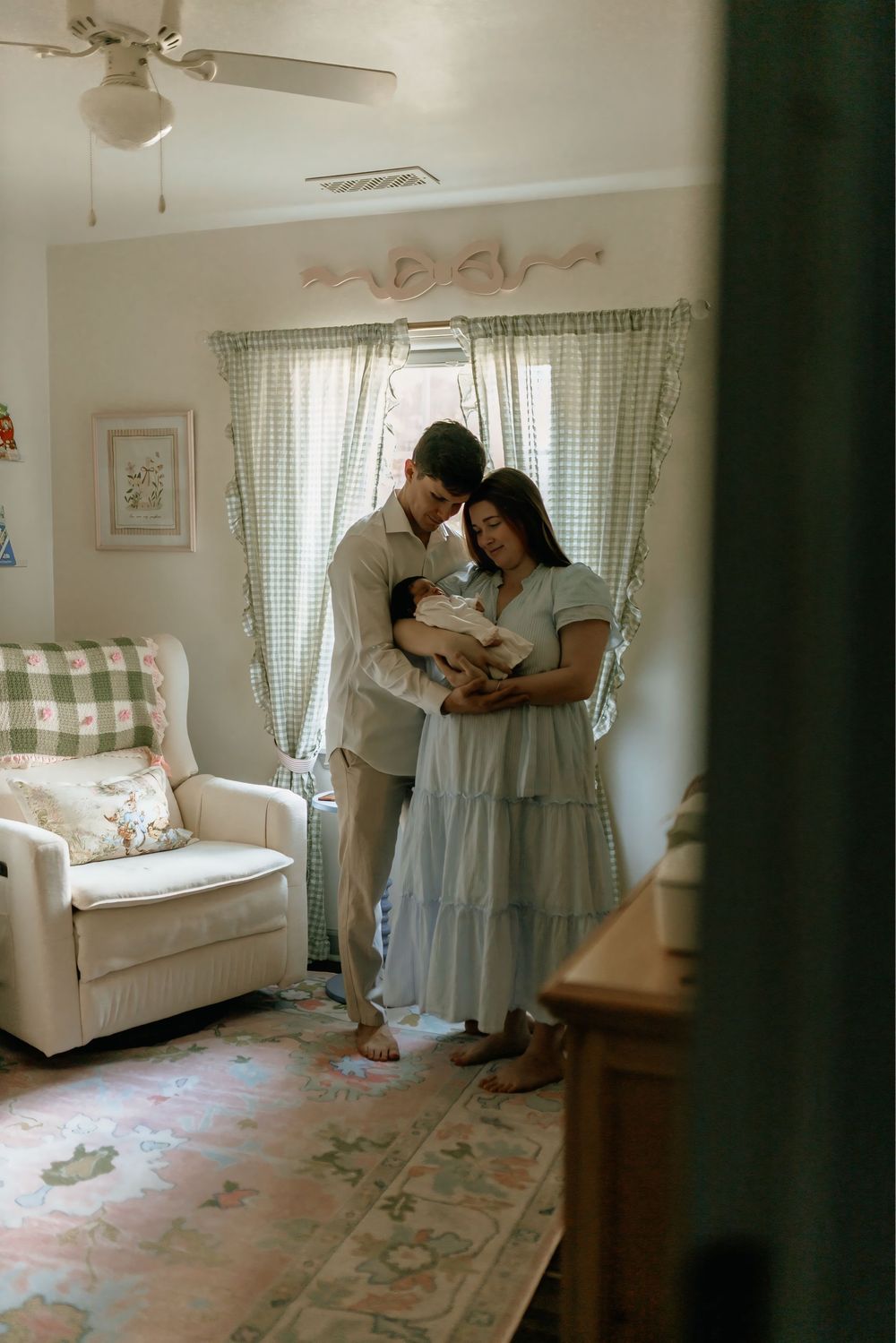 New parents stand together in a soft nursery, tenderly cradling their newborn baby in a cozy, pastel-decorated room.