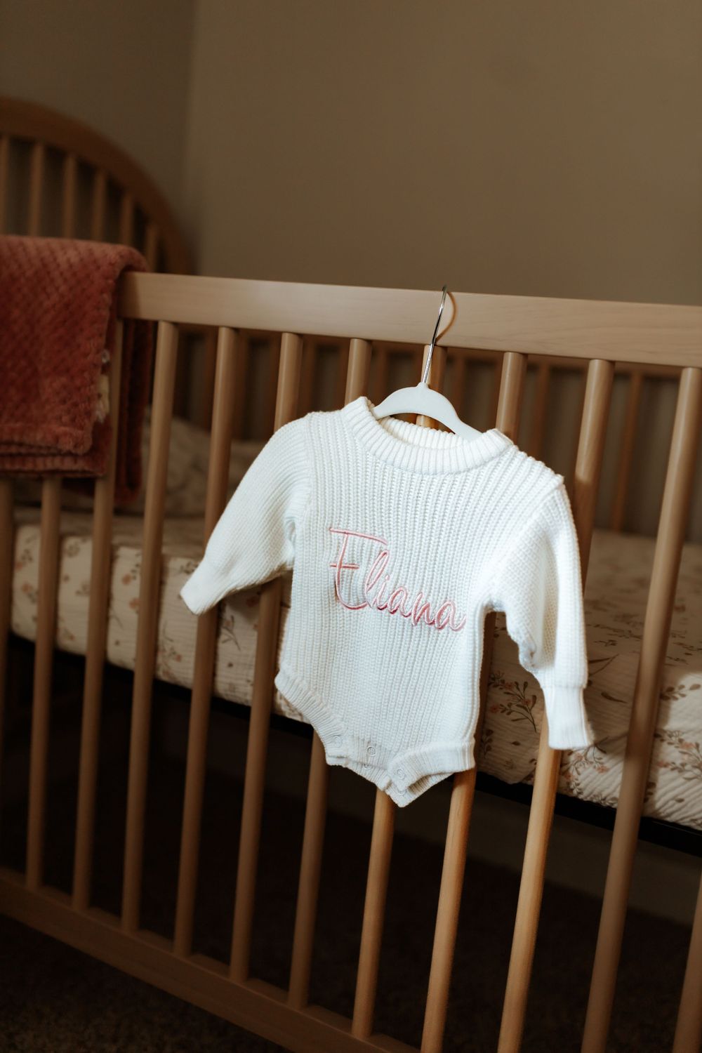 White ribbed baby onesie with pink embroidery hangs on wooden crib in nursery setting.