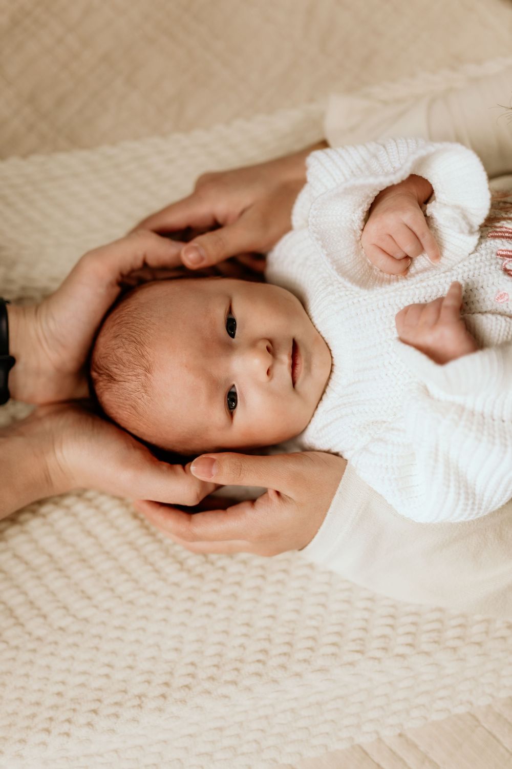 A tiny baby wearing a white knit sweater lies on a textured cream blanket while gentle hands cradle their head.
