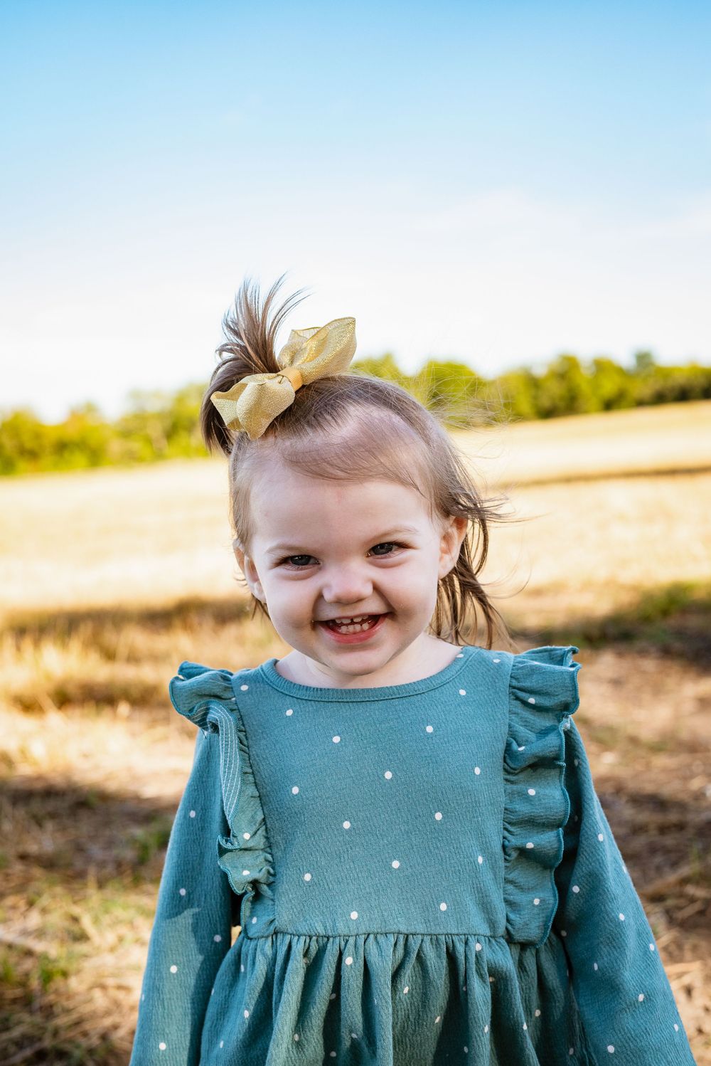 Toddler wearing teal ruffled dress smiles outdoors in golden sunlight.