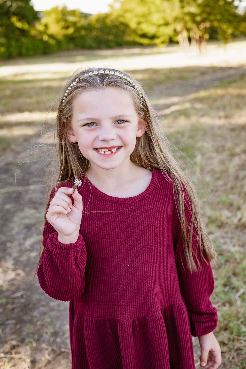 Child in burgundy sweater dress poses outdoors with natural backdrop.