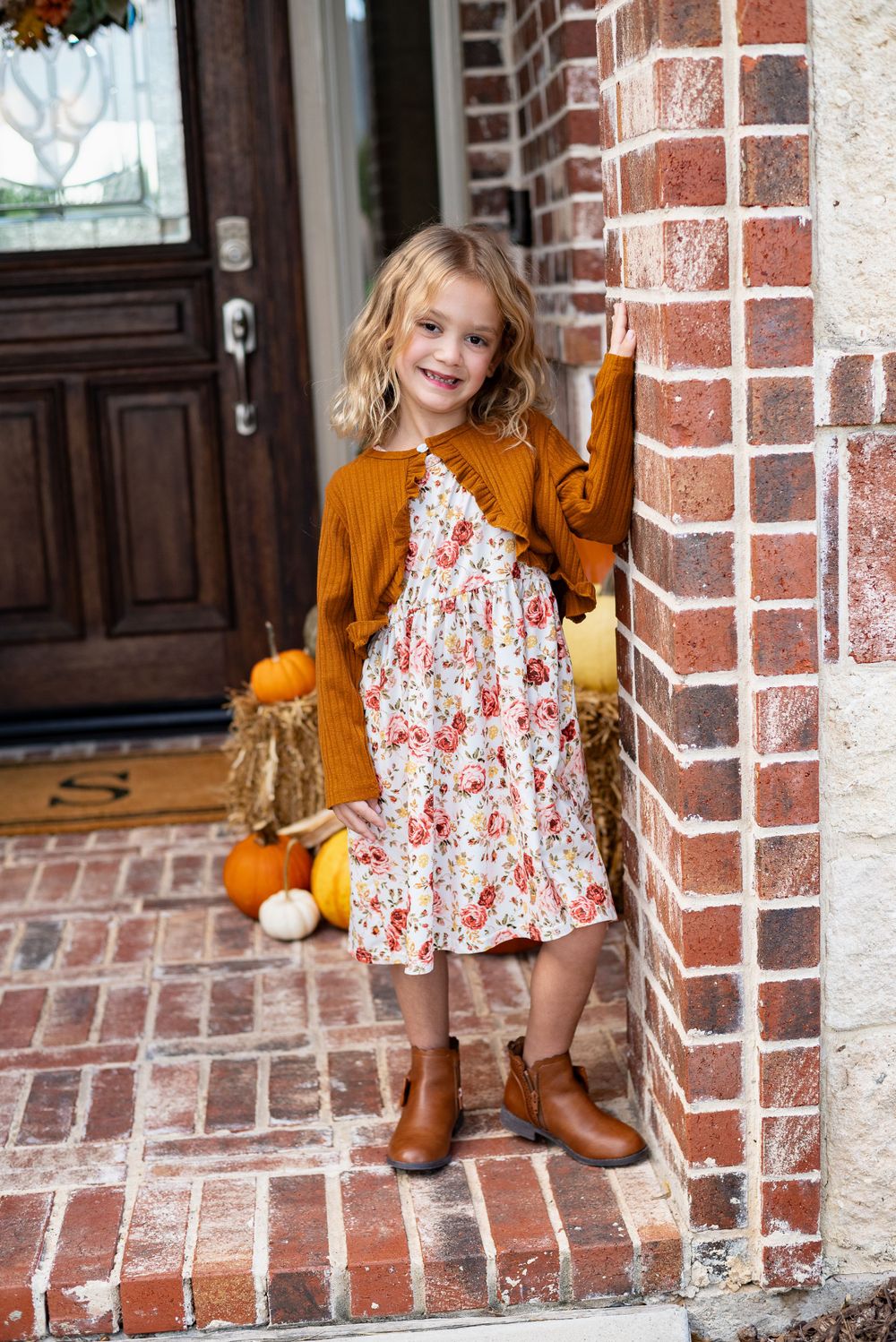 Child in floral dress and mustard cardigan poses on brick porch with fall decorations.