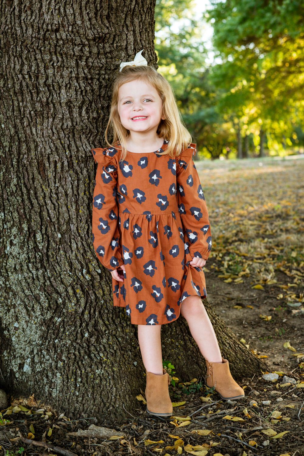 Child in orange floral dress poses by tree trunk with fallen autumn leaves.