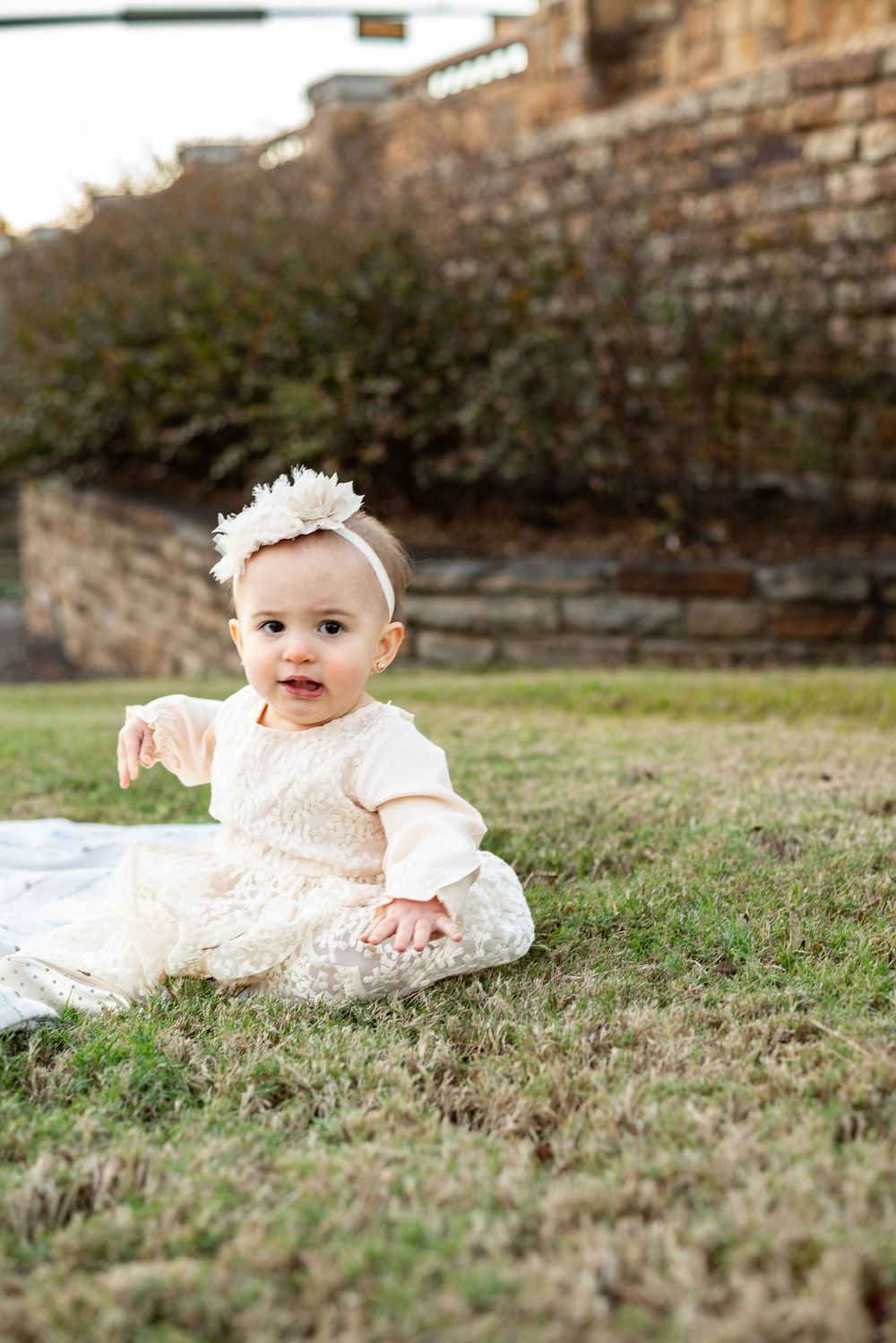 Baby in cream lace dress sits on grass near stone wall background.