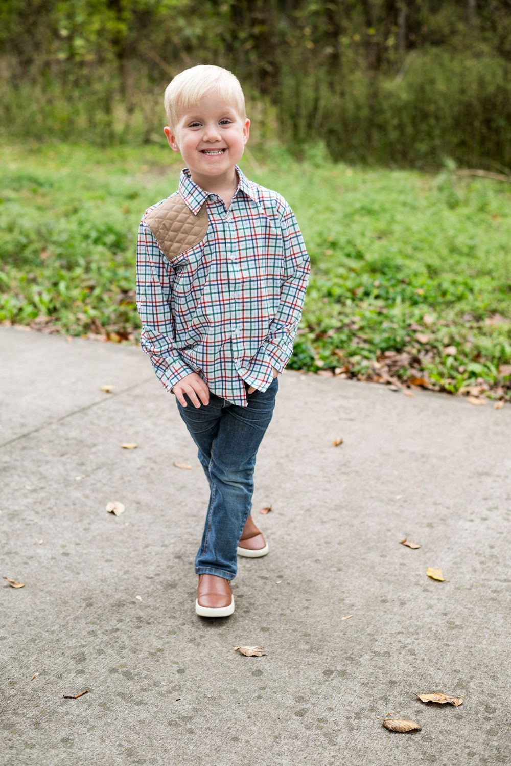 Child in plaid shirt and jeans standing on outdoor path with greenery in background.