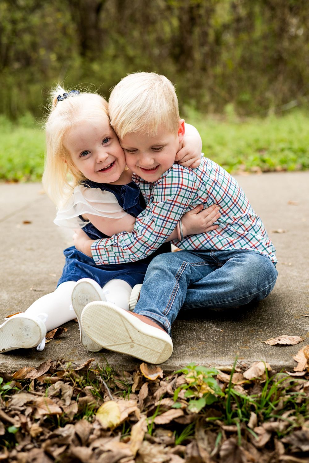 Two young children hugging and laughing while sitting on concrete path with autumn leaves.