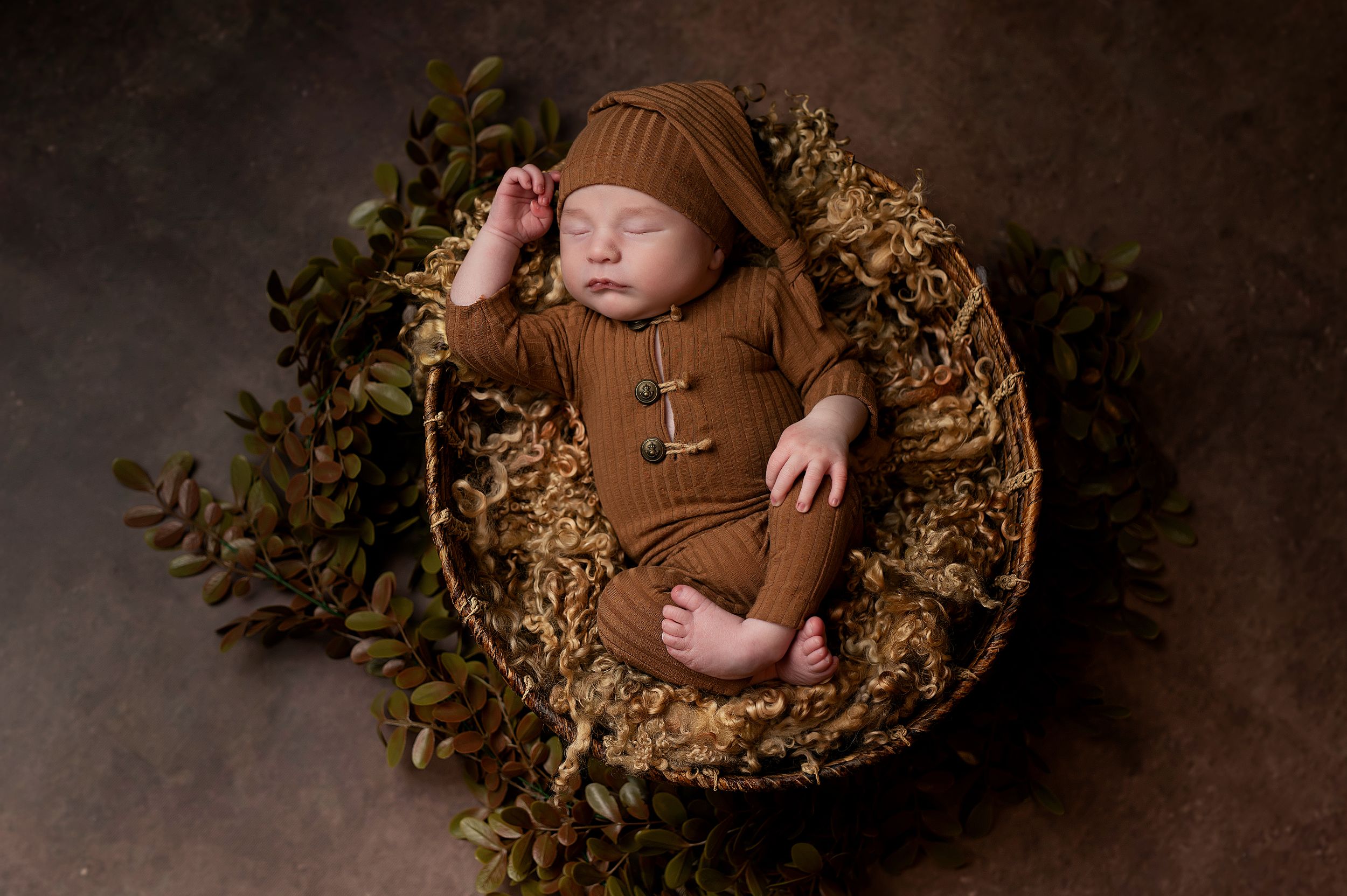 A newborn baby sleeps peacefully in a rustic brown nest surrounded by autumn leaves and branches in a dark setting.